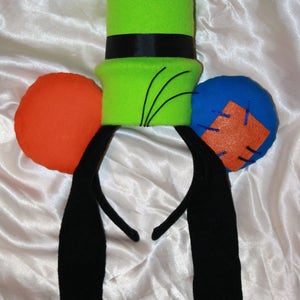 Puede incluir: Una diadema de felpa inspirada en Goofy, con un sombrero verde brillante con una banda negra, orejas negras y detalles circulares naranjas y azules. La diadema está diseñada para parecerse al aspecto icónico del personaje de dibujos animados.
