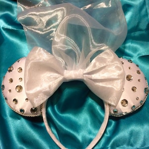 Può includere: Orecchie di Minnie Mouse in raso bianco con un grande fiocco bianco e un velo di tulle bianco. Le orecchie sono decorate con strass trasparenti.