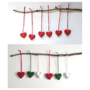 May include: A display of heart-shaped decorations in red, green, and white, hanging from a natural branch. The ornaments are crafted from a textured fabric and are suspended by red, green, and white cords. The backdrop is a simple white surface.