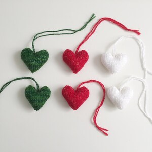May include: Six knitted heart ornaments in green, red, and white, each with a matching string for hanging. The hearts are arranged on a white surface, with a close-up view highlighting the texture and colors.