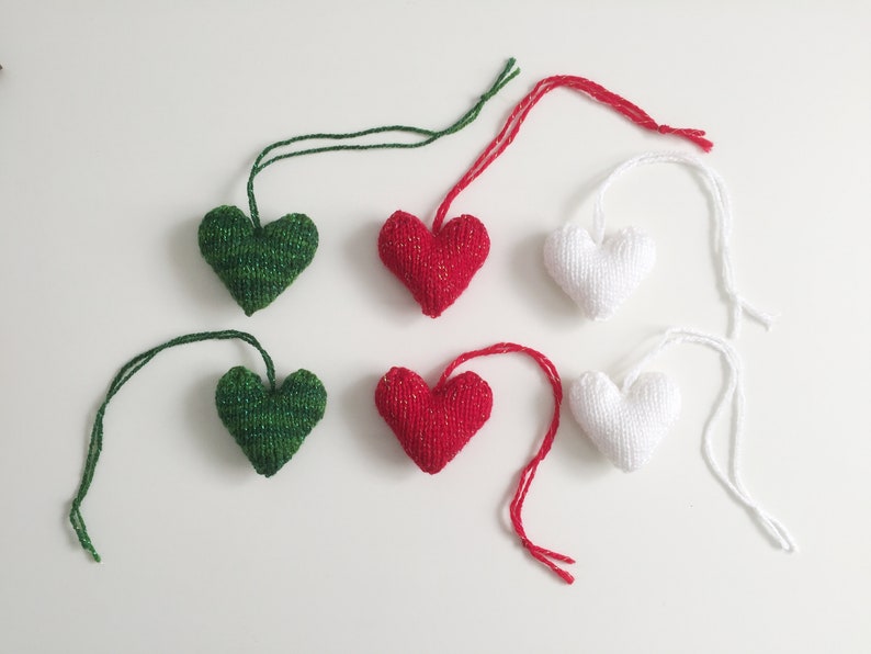 May include: Six knitted heart ornaments in green, red, and white, each with a matching string for hanging. The hearts are arranged on a white surface, with a close-up view highlighting the texture and colors.