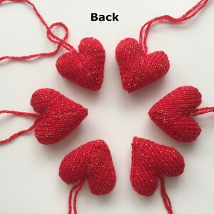 May include: Six red knitted heart ornaments with gold flecks are arranged in a circle. Each heart has a red string attached for hanging. The word "Back" is visible in the image. These ornaments are suitable for Valentine's Day or other celebrations.