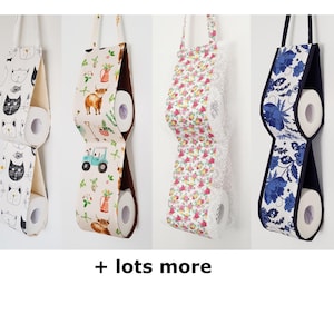 May include: Four hanging fabric toilet paper holders with loops for wall mounting. Each holder features two stacked rolls of toilet paper. Patterns include black and white cat faces, farm animals with a tractor, pink and green floral, and blue floral. + lots more