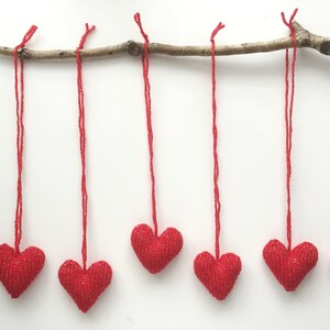 May include: Six red heart-shaped ornaments hang from a natural branch against a white background. Each heart is suspended by a red string. The hearts have a textured, glittery appearance, suggesting a festive or decorative purpose.