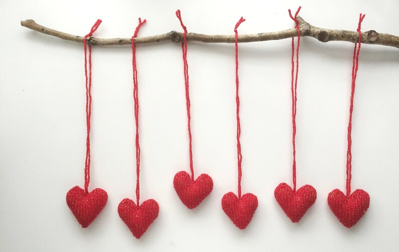 May include: Six red heart-shaped ornaments hang from a natural branch against a white background. Each heart is suspended by a red string. The hearts have a textured, glittery appearance, suggesting a festive or decorative purpose.