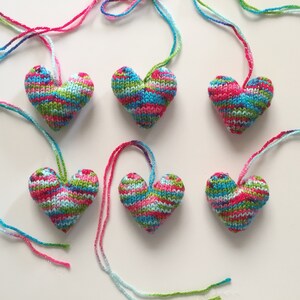 May include: Six knitted heart ornaments in shades of pink, blue, green, and white. Each heart has a colorful yarn loop for hanging. The hearts are arranged on a white surface.