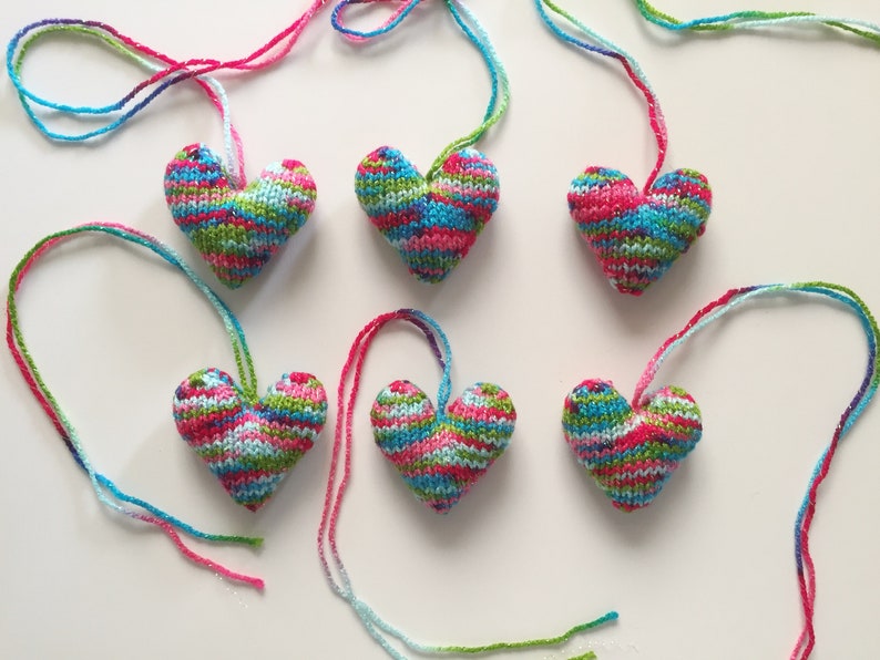 May include: Six knitted heart ornaments in shades of pink, blue, green, and white. Each heart has a colorful yarn loop for hanging. The hearts are arranged on a white surface.