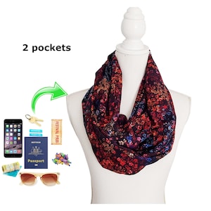 May include: A floral infinity scarf in shades of burgundy, red, and blue is displayed on a white mannequin. The scarf has two pockets. Accessories such as a phone, keys, passport, sunglasses, and a festival pass are shown.