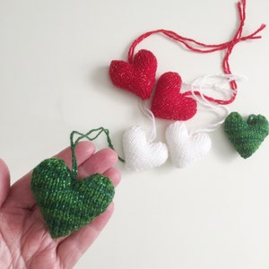 May include: A collection of knitted heart ornaments in red, white, and green. The red hearts have a gold shimmer, while the green heart has a glittery effect. Each heart has a string for hanging, perfect for Valentine's Day or other celebrations.