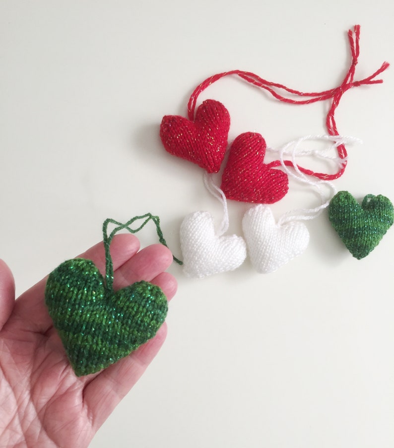 May include: A collection of knitted heart ornaments in red, white, and green. The red hearts have a gold shimmer, while the green heart has a glittery effect. Each heart has a string for hanging, perfect for Valentine's Day or other celebrations.
