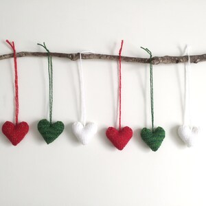 May include: Six knitted heart ornaments in red, green, and white hang from a natural branch. Each heart is suspended by a matching colored string. The ornaments are arranged against a white background.