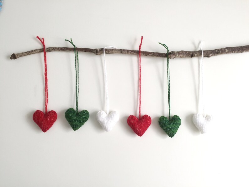 May include: Six knitted heart ornaments in red, green, and white hang from a natural branch. Each heart is suspended by a matching colored string. The ornaments are arranged against a white background.