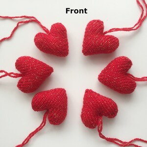 May include: Six red knitted heart ornaments with gold metallic thread accents. Each heart has a string attached for hanging. The hearts are arranged in a circular pattern against a white background.