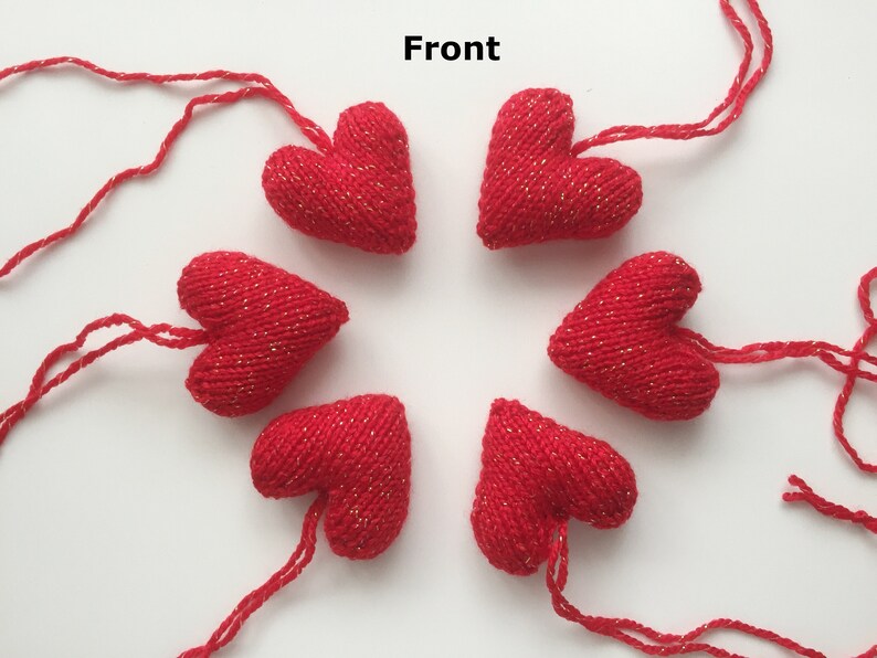 May include: Six red knitted heart ornaments with gold metallic thread accents. Each heart has a string attached for hanging. The hearts are arranged in a circular pattern against a white background.