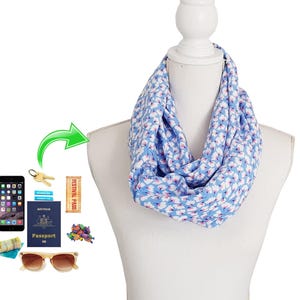 May include: A blue infinity scarf with a white floral pattern is displayed on a mannequin. The scarf is paired with travel accessories, including a smartphone, keys, passport, festival pass, sunglasses, and money.