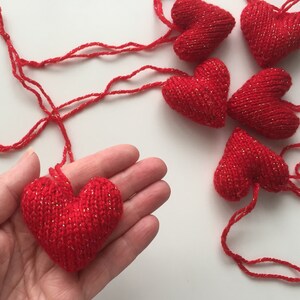 May include: Red knitted heart ornaments with gold metallic thread accents. One heart is held in a hand, with several others scattered on a white surface. These handmade hearts are perfect for Valentine's Day or other special occasions.