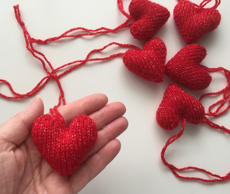 May include: Red knitted heart ornaments with gold metallic thread accents. One heart is held in a hand, with several others scattered on a white surface. These handmade hearts are perfect for Valentine's Day or other special occasions.