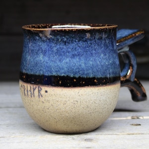 May include: A handmade ceramic mug with a unique handle. The mug features a speckled beige base, a dark band, and a gradient of blue hues. The rim and handle are dark brown. The word "RHRYR" is inscribed on the side.