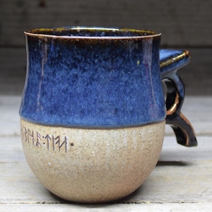 May include: A blue and beige ceramic mug with a brown handle. The mug has a dark blue glaze on the top half and a beige glaze on the bottom half. The bottom half of the mug has a line of black runes.