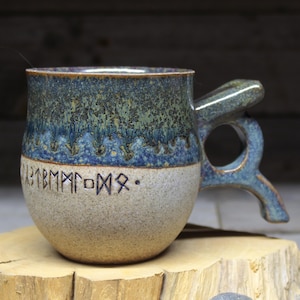 May include: A handcrafted ceramic mug with a unique handle design. The mug features a two-tone glaze, with a blue and green top and a tan base. Runes are etched around the base of the mug. The mug is displayed on a wooden slice.
