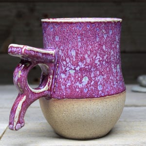May include: A handmade ceramic mug with a unique, asymmetrical handle. The mug is glazed in a vibrant pink with white speckles and a beige bottom.