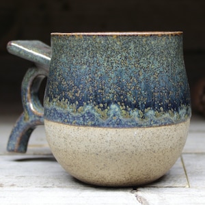 May include: A handcrafted ceramic mug with a distinctive handle. The mug has a speckled blue and teal glaze over a tan base. The rim has a brown accent. The mug is designed for hot drinks.