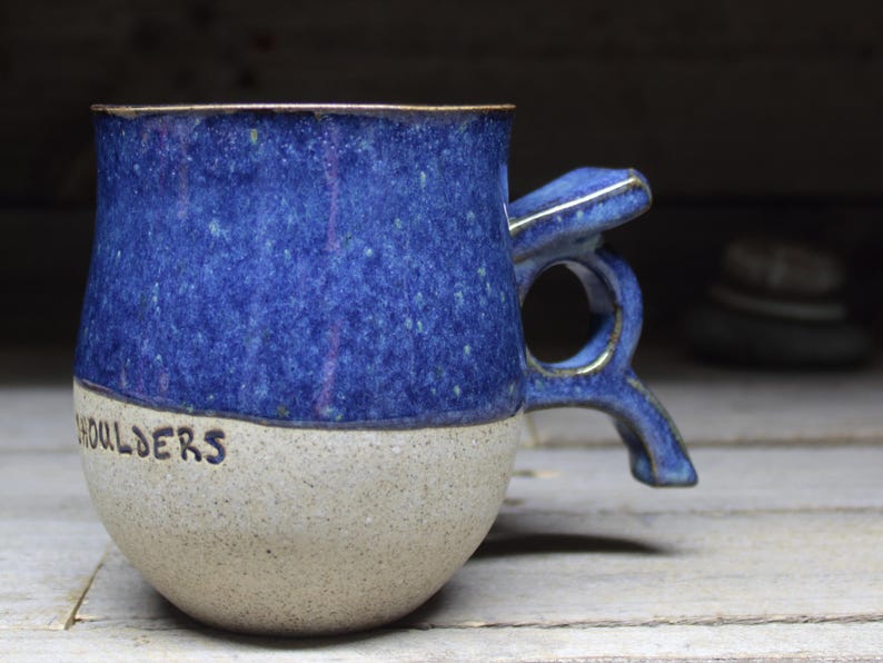 May include: A handmade ceramic mug with a blue speckled glaze on the upper half and a beige speckled base. The word "SHOULDERS" is written in black on the base. The handle has a unique, sculptural design.