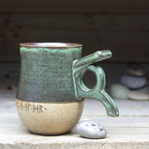 May include: A green and brown ceramic mug with a unique handle. The mug has a light brown base with runes inscribed on it.