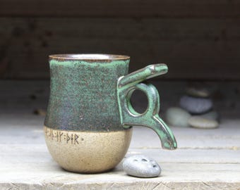 May include: A green and brown ceramic mug with a unique handle. The mug has a light brown base with runes inscribed on it.