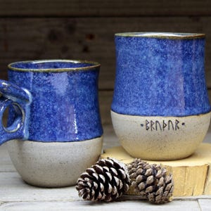 May include: Two handmade ceramic mugs with blue and white speckled glaze. One mug has a unique handle and the other has the word "BRNBR" written on the bottom in black.