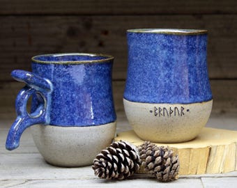 May include: Two handmade ceramic mugs with blue and white speckled glaze. One mug has a unique handle and the other has the word "BRNBR" written on the bottom in black.