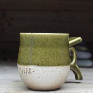 May include: A handmade ceramic mug with a unique handle. The top half is a mottled olive green, while the bottom is a speckled off-white. The word "Yggdrasil" is etched in runes near the base. A rustic, earthy design.