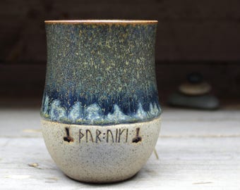 May include: A ceramic mug with a blue and green glaze and a white rim. The mug has a inscription in runes that reads "FARK-NIRI".