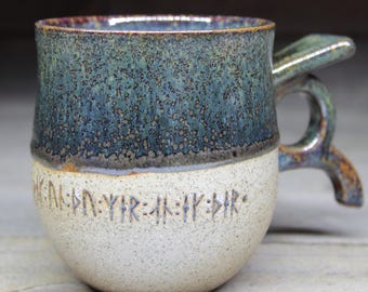 May include: Handmade ceramic mug with a unique handle design. The top half is a speckled blue-green glaze, contrasting with the off-white base. The base features etched text. A rustic, artisanal piece.