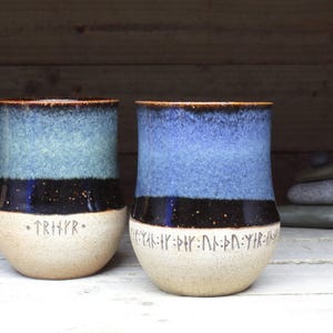 May include: Two ceramic mugs with blue and black glaze. The mugs have a white rim and base. The mugs have runes inscribed on the bottom.