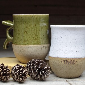 May include: Two ceramic mugs with a speckled glaze. The mug on the left is green with a white rim and a brown handle. The mug on the right is white with a brown rim. Both mugs have the word "Yggdrasil" written on the bottom in brown.