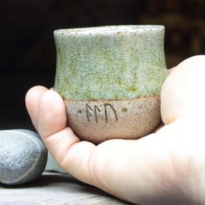 Puede incluir: Una taza pequeña de cerámica verde y marrón con un esmalte blanco y un símbolo grabado en el lateral. La taza está siendo sostenida en una mano.