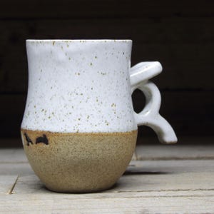 May include: A white ceramic mug with a speckled glaze and a brown rim. The mug has a unique handle that is shaped like a loop.