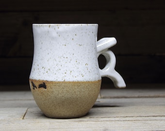 May include: A white ceramic mug with a speckled glaze and a brown rim. The mug has a unique handle that is shaped like a loop.