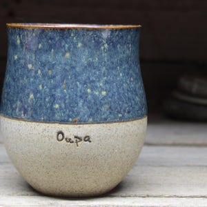 May include: A handmade ceramic tumbler with a speckled blue glaze on the upper half and a light beige base. The word "Oupa" is inscribed on the base. The rim has a brown accent.