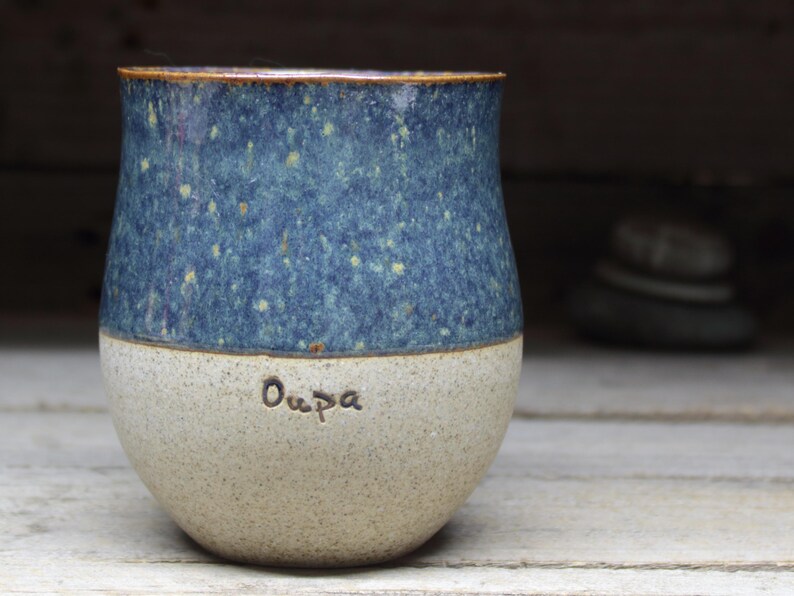 May include: A handmade ceramic tumbler with a speckled blue glaze on the upper half and a light beige base. The word "Oupa" is inscribed on the base. The rim has a brown accent.