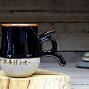 May include: A black and white ceramic mug with a unique handle. The mug has a black glaze and a white base with black runes that read "May your path be clear".