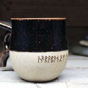 May include: A handcrafted ceramic mug with a black speckled top and a speckled beige bottom. The mug features a dark brown handle and rim. Runes are etched into the beige portion of the mug.