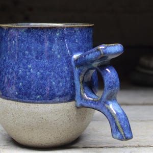 May include: A handmade ceramic mug with a unique handle design. The upper portion of the mug is a speckled blue, while the lower half is a speckled beige. The rim has a gold accent. The handle is integrated into the design, creating a distinctive look.