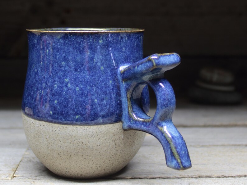 May include: A handmade ceramic mug with a unique handle design. The upper portion of the mug is a speckled blue, while the lower half is a speckled beige. The rim has a gold accent. The handle is integrated into the design, creating a distinctive look.