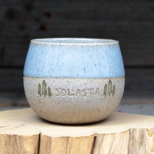 Könnte beinhalten: Eine hellblaue und weiße Keramiktasse mit dem Wort "SOLASTA" und einem Design aus drei Bäumen an der Seite.