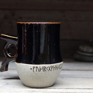 May include: A black and white ceramic mug with a handle shaped like a stylized arm. The mug has a black glaze on the top half and a white glaze on the bottom half. The bottom half has black runes inscribed on it.