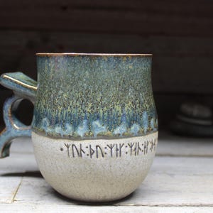 Könnte beinhalten: Handgefertigte Keramiktasse mit einzigartigem Griff und zweifarbigem Glaze. Die obere Hälfte ist ein meliertes Türkis und Blau, während die Unterseite hellbeige ist. Runen sind um den Boden der Tasse herum geätzt.