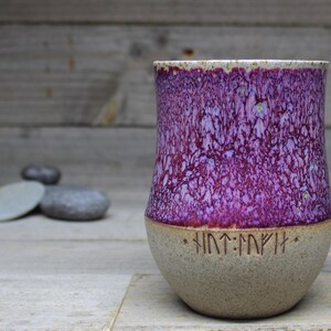 May include: A ceramic mug with a purple and white speckled glaze. The mug has a rounded bottom and a wide mouth. The bottom of the mug has a row of runes.