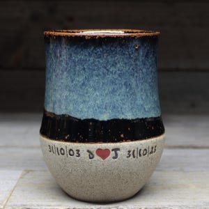 May include: A blue and black ceramic mug with a white rim and base. The mug has a heart with the letter "J" inside and the date "3/11/03" on one side and the date "3/11/03" on the other side.